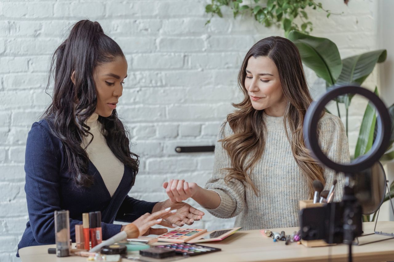 Jeunes blogueuses positives et multiethniques essayant des produits de maquillage tout en étant assises à une table et en enregistrant un vlog sur un smartphone posé sur un trépied avec ring light